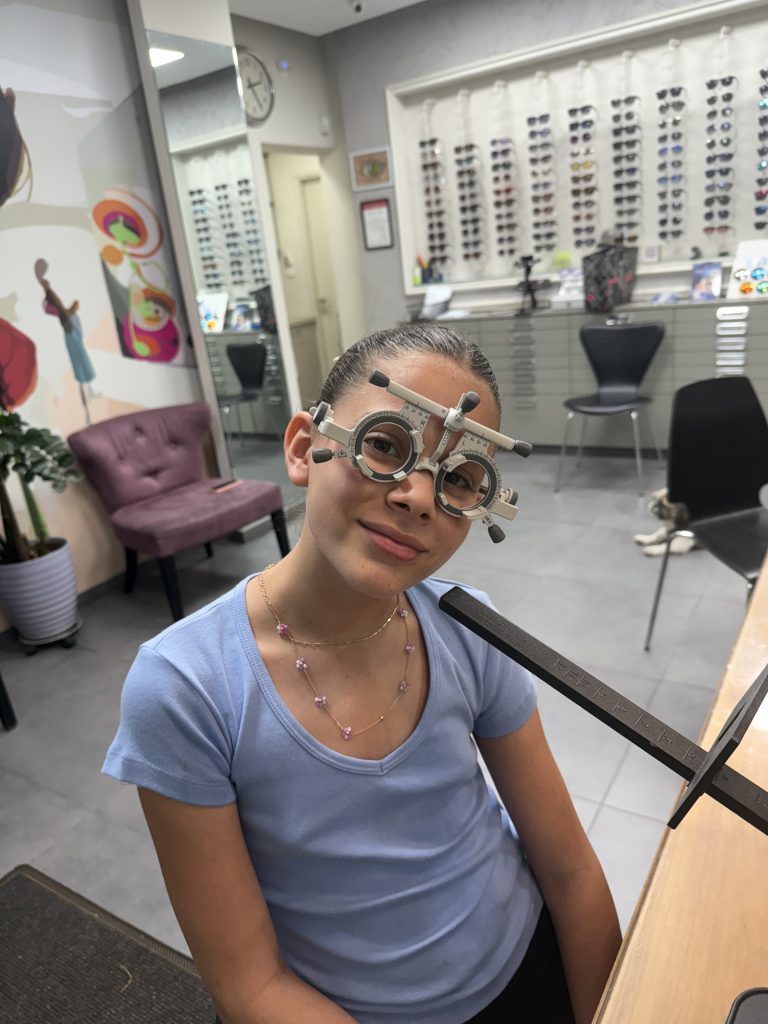 ילדה עם משקפי ניסיון בבדיקת ראייה לילדים מול קיר המסגרות באופטיטופ